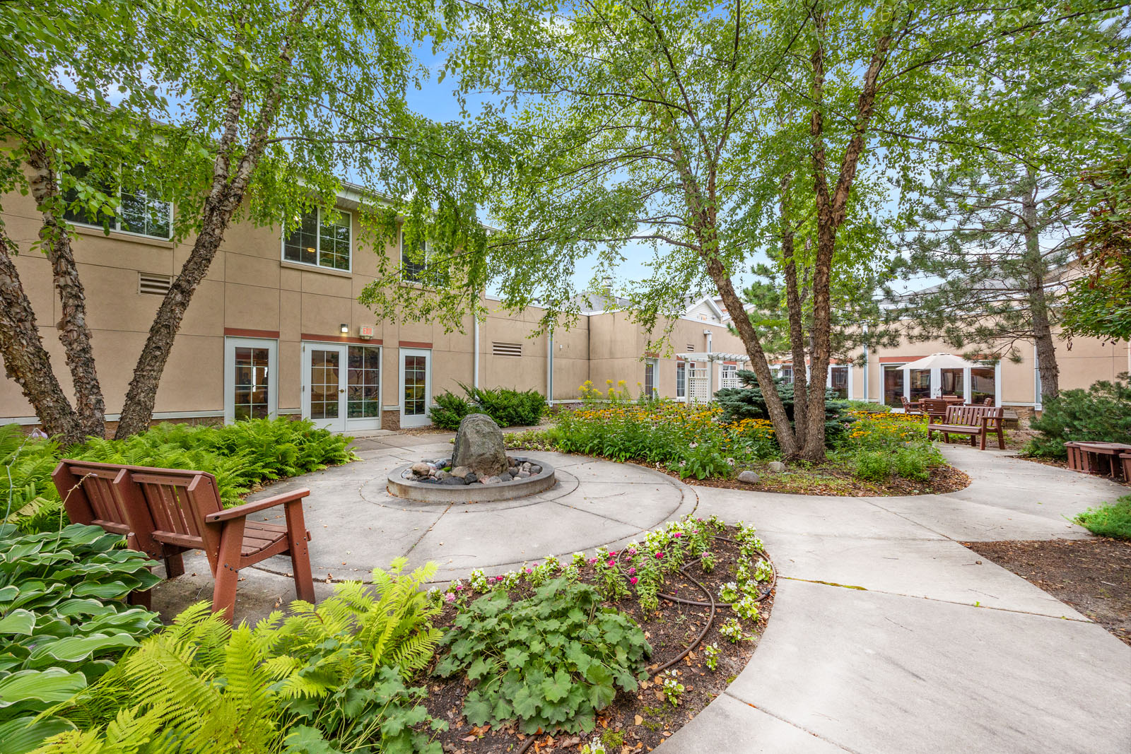 A landscaped courtyard with flowers, a fountain, benches, trees, and a beige building in the background.