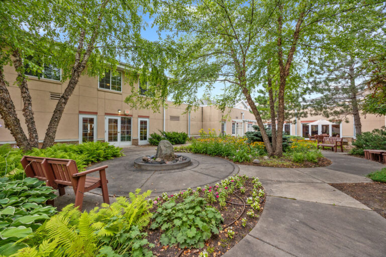A landscaped courtyard with flowers, a fountain, benches, trees, and a beige building in the background.