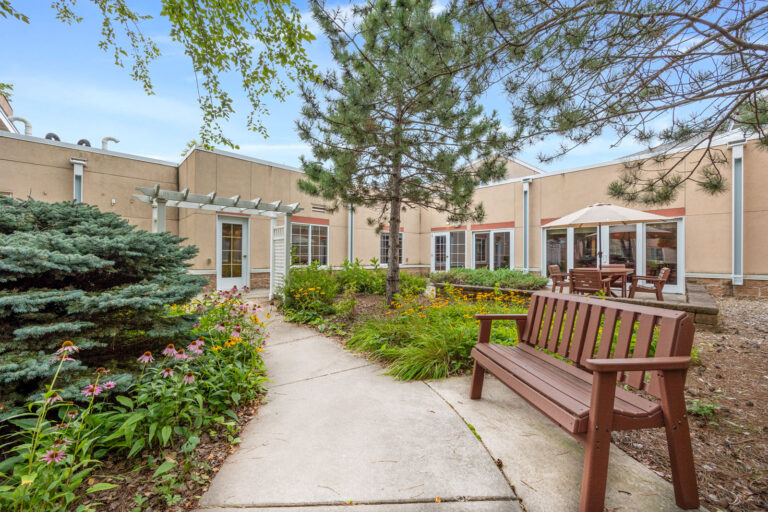 A garden courtyard with benches, flowers, trees, and patio tables outside a beige building.