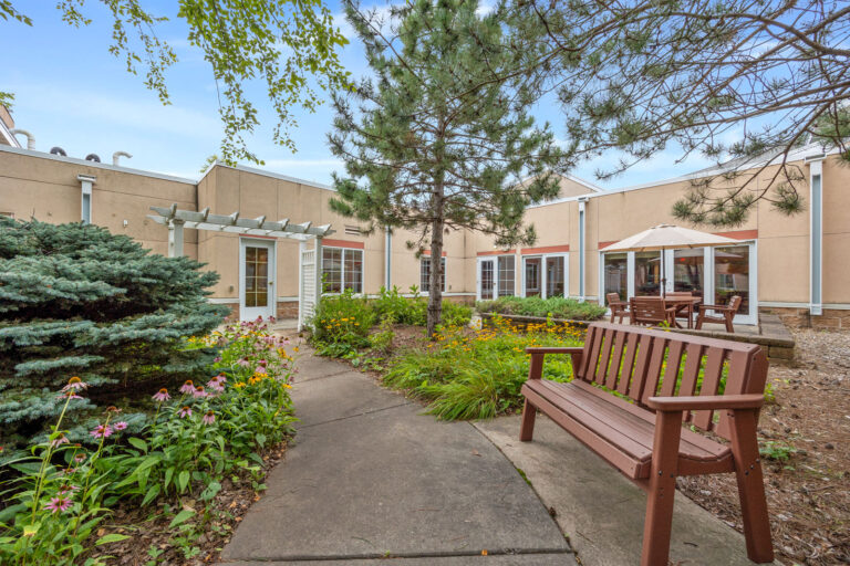 A garden courtyard with benches, flowers, trees, and patio tables outside a beige building.