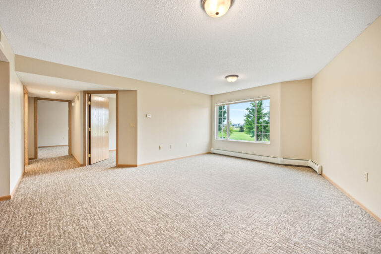 Empty apartment living room with carpet flooring, large window, and doorway leading to a hallway and other rooms.