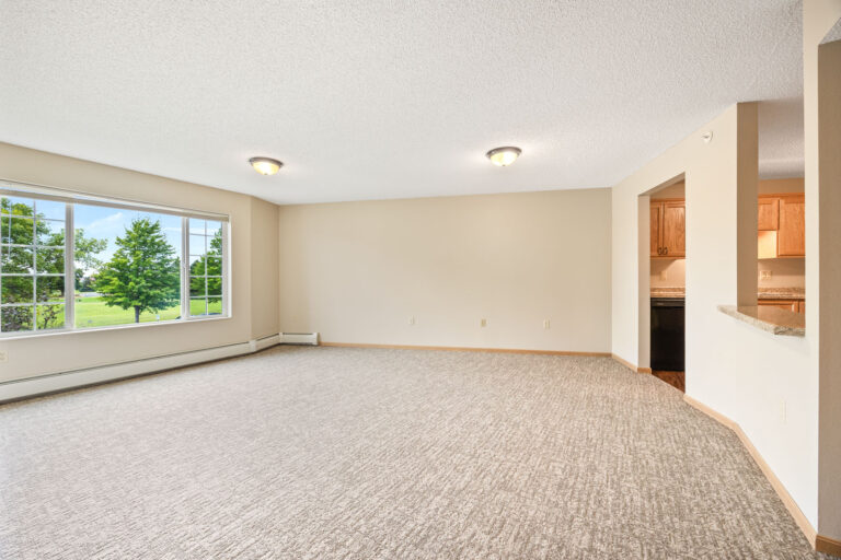Spacious, empty apartment living room with carpet, large window, beige walls, and view into a kitchen.