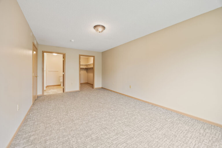 Empty apartment bedroom with carpet, ceiling light, open door to bathroom, and walk-in closet.