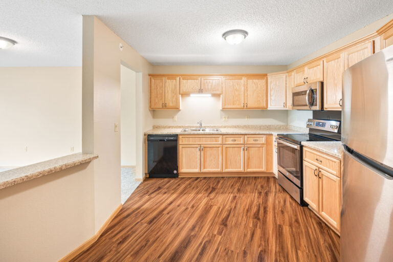 Modern independent living apartment kitchen with wooden cabinets, stainless steel appliances, and wood-style flooring.