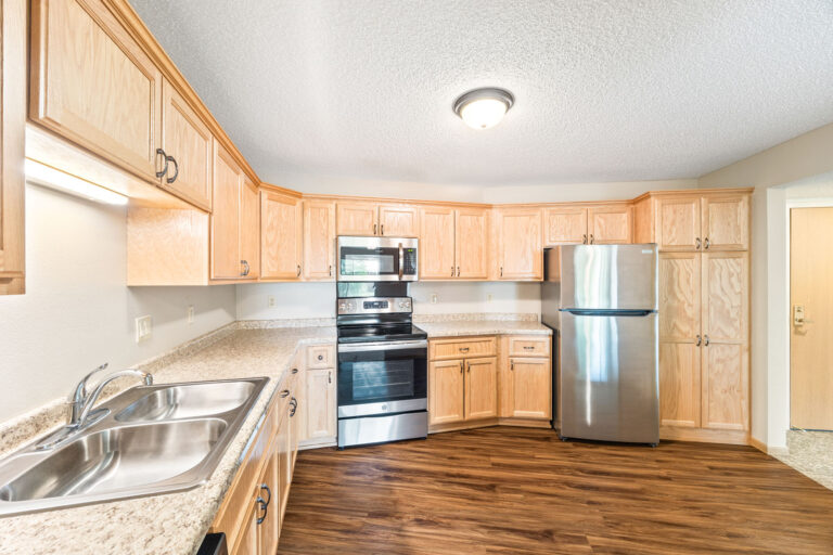 Modern kitchen with light wood cabinets, stainless steel appliances, and a double sink on granite counters.