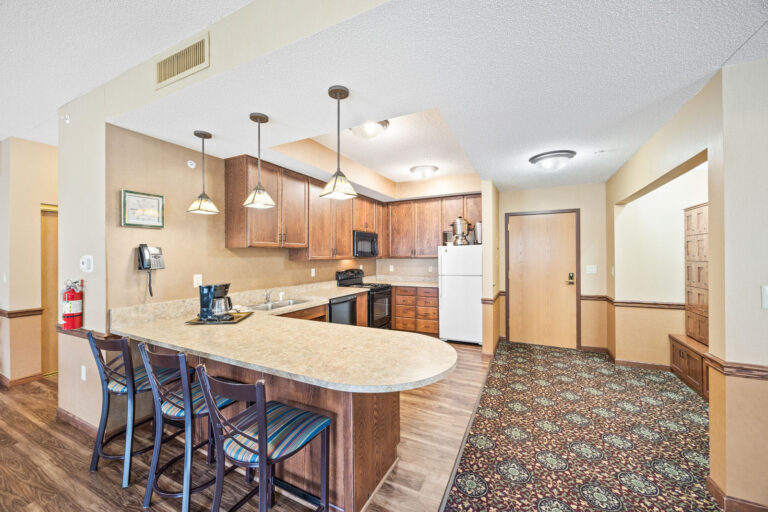 Independent Living spacious hotel kitchen with bar seating, wooden cabinets, appliances, and patterned carpet in entryway.