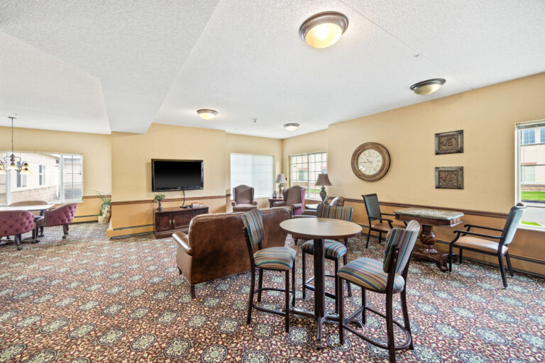 Bright lounge with patterned carpet, chairs, tables, a TV, and large windows letting in natural light.
