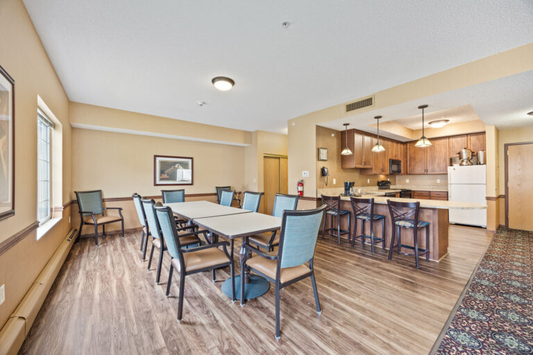 Clubroom dining area with tables and chairs next to a kitchen with wooden cabinets and barstools.