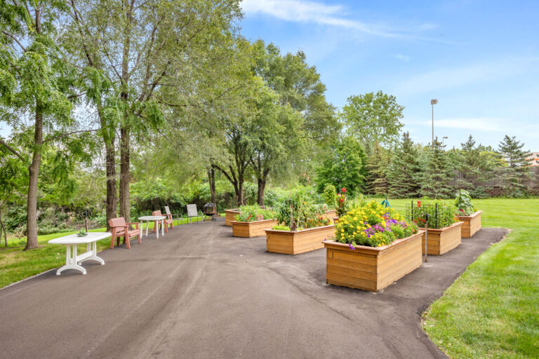 Wooden raised garden beds with colorful flowers and plants beside outdoor seating on a paved path in a park.