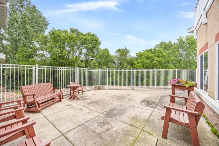Spacious patio with wooden benches, planter flowers, and white metal fence, surrounded by lush green trees.