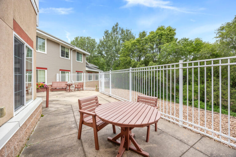 Memory care outdoor patio with wooden tables and chairs, surrounded by a white metal fence and greenery.