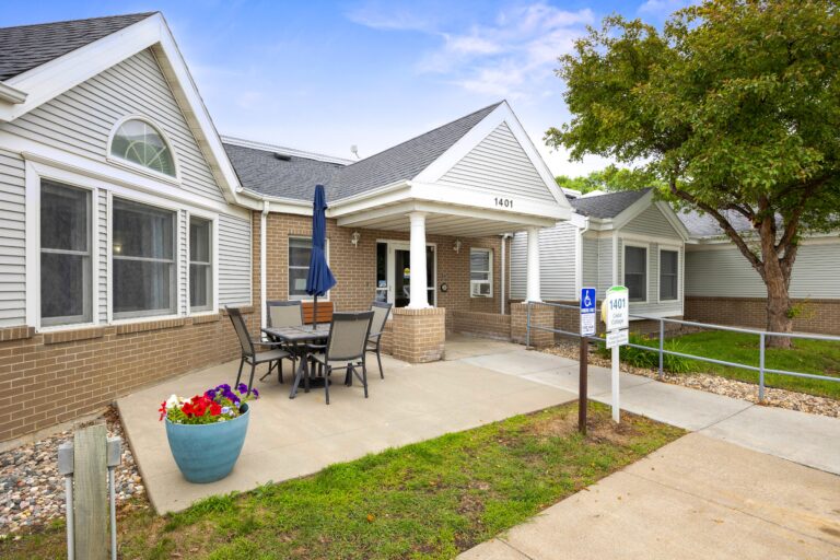Single-story basic care 1401 building entrance with patio, outdoor seating, blue umbrella, and large potted plant.