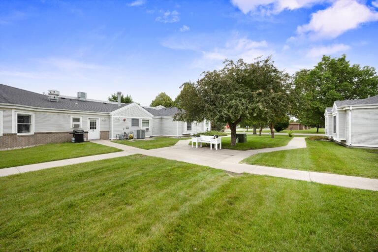 Single-story white buildings surround a green courtyard with trees and a picnic table under the sun.