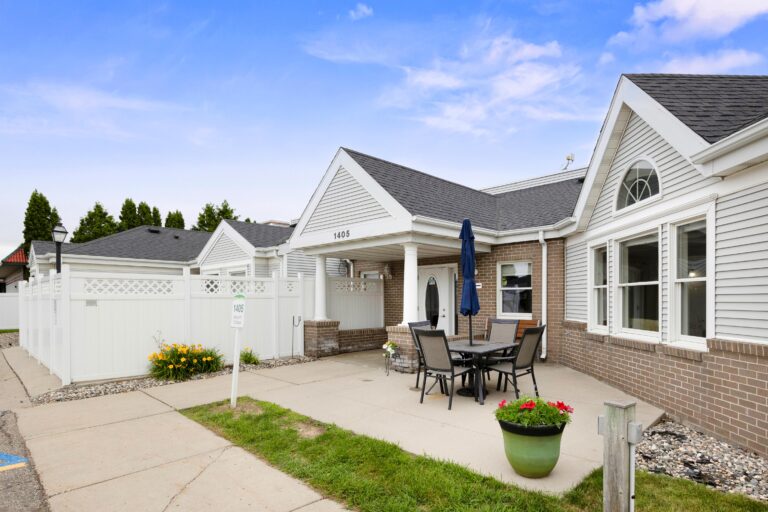 Single-story memory care 1405 building entrance with patio table, umbrella, flowers, and white fence.
