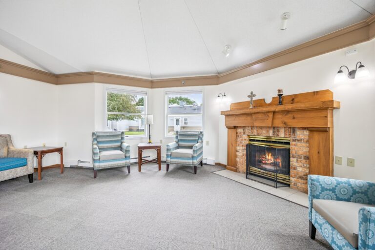 A cozy memory care living room with striped chairs, a brick fireplace, and large windows letting in natural light.