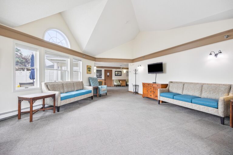 Spacious memory care living room with sofas, blue chairs, a TV, large window, and neutral décor.