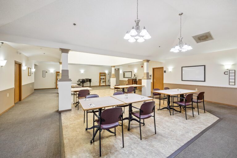 Bright, spacious memory care dining room with tables, chairs, and overhead lights in a modern building.