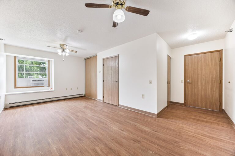 Bright empty apartment with wood flooring, ceiling fans, window, and wooden doors.