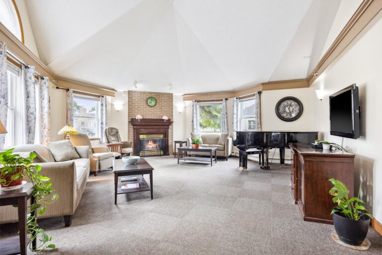 Bright basic care living room with fireplace, sofa, grand piano, TV, and large windows letting in natural light.