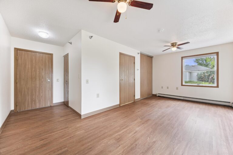 Empty apartment with wood floors, white walls, wooden doors, ceiling fans, and a window with outdoor view.