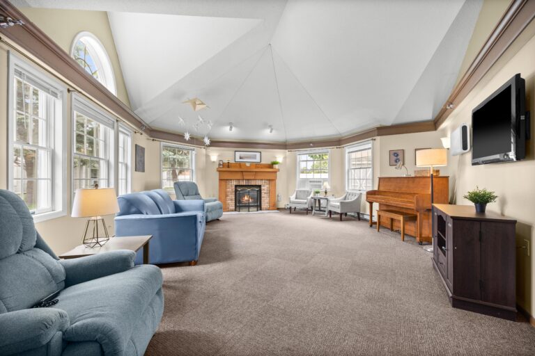 Spacious living room with blue chairs, a fireplace, piano, TV, and large windows letting in natural light.
