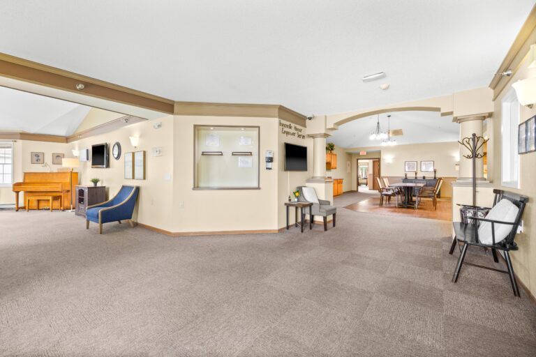 Spacious, carpeted common area with seating, a piano, and a dining room in the background.