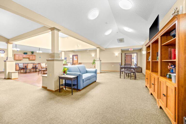 A bright, open lounge with a blue sofa, dining tables, and a wooden bookshelf in a communal space.