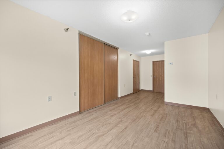 Empty apartment with beige walls, wood floor, and wooden doors; ceiling lights are on.