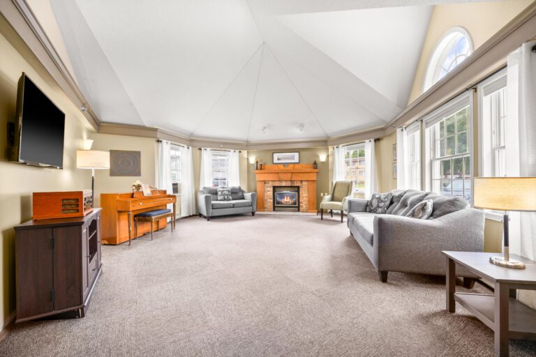 Bright living room with vaulted ceiling, fireplace, sofas, piano, and large windows letting in natural light.