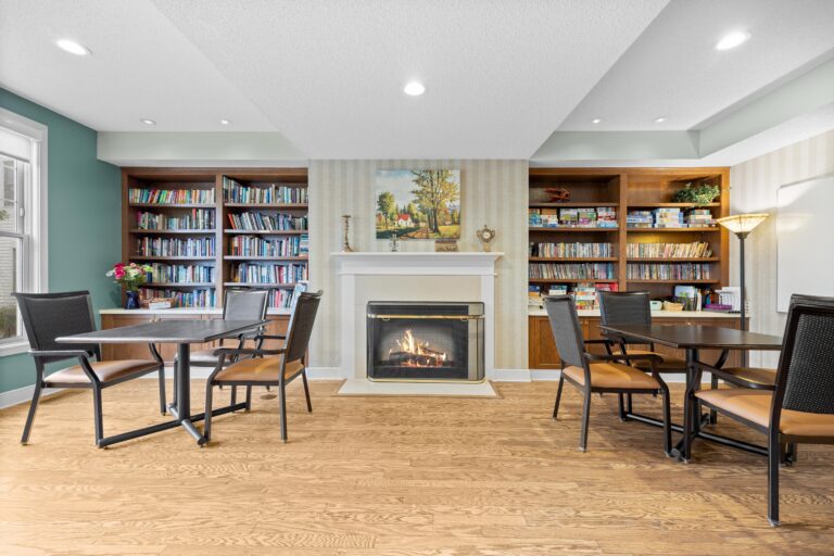 A cozy library room with bookshelves, a fireplace, tables, and chairs for reading or study.