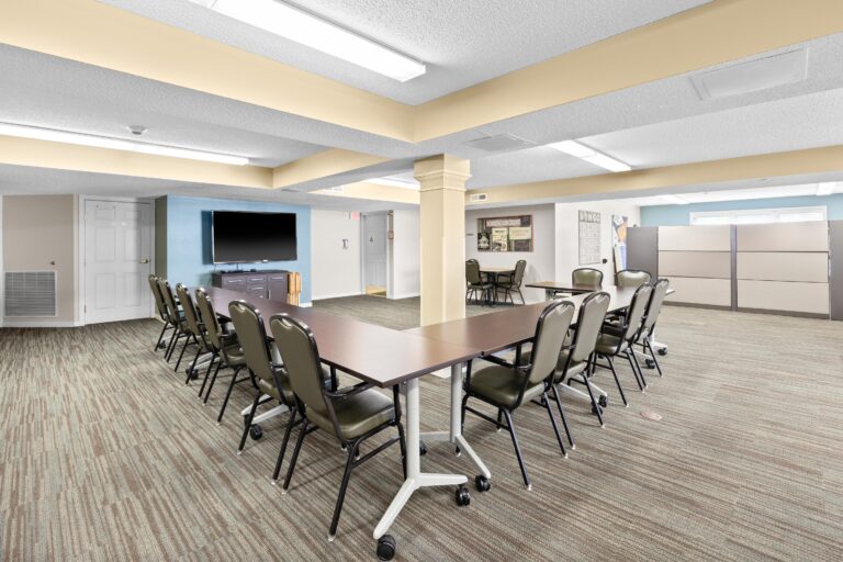 Large meeting room with tables, chairs on wheels, TV, and carpeted floor under fluorescent lights.