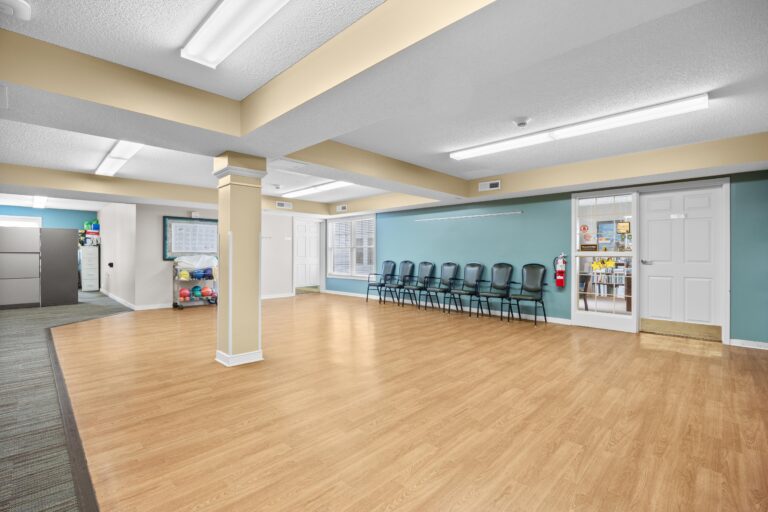 Spacious room with wood floor, blue wall, a row of black chairs, and office area visible through glass door.