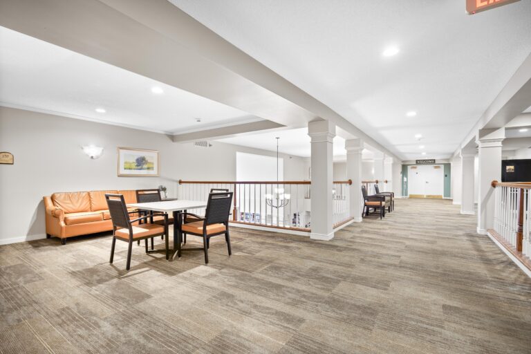 Bright hallway with carpet, table and chairs, armchair, white railings, and wall art in a modern building.