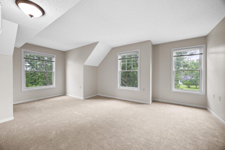 Bright empty apartment room with beige carpet, three large windows, and neutral-colored walls.