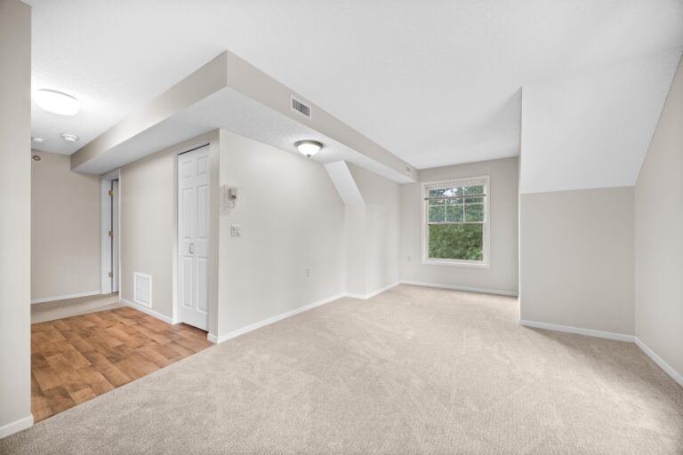 Bright, empty apartment room with beige carpet, white walls, a large window, and a small entryway with wood flooring.