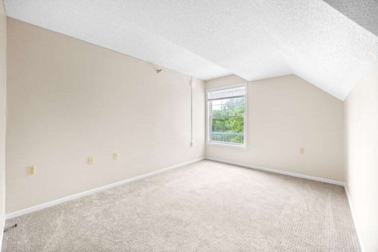 Empty beige apartment bedroom with carpeted floor, off-white walls, a single window, and sloped ceiling.