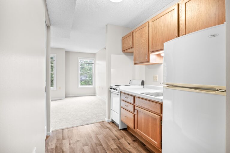 Small apartment kitchen with wooden cabinets, white appliances, and a view into a bright, carpeted living area.
