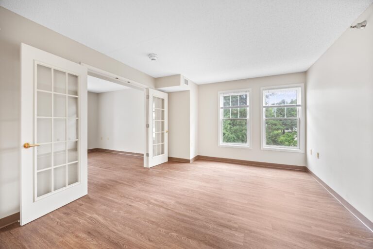 Bright empty room with wood flooring, French doors, and two large windows overlooking greenery.