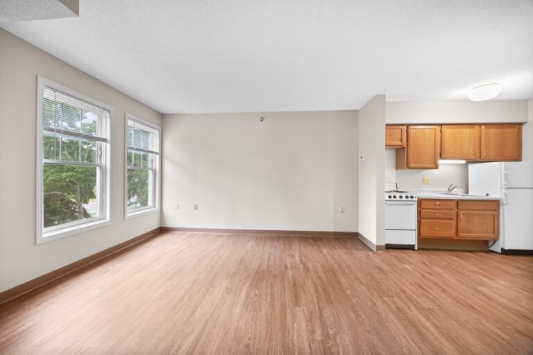 Bright, empty apartment room with wood floors, large windows, and a small kitchen area with wood cabinets.