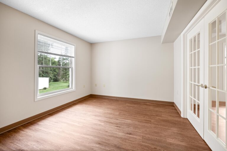 Empty room with wood floors, beige walls, a large window, and glass double doors letting in natural light.