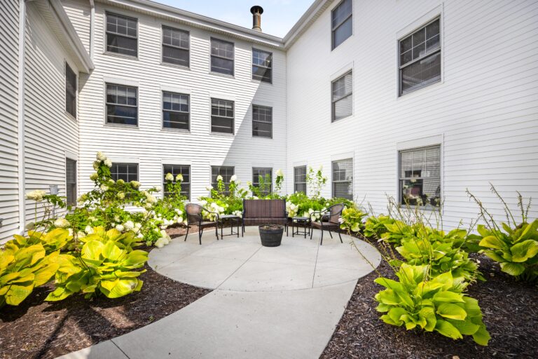 Small courtyard with green plants, outdoor seating, and white building walls with windows surrounding the area.
