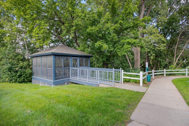 A blue screened gazebo with a ramp, surrounded by green grass and trees next to a paved walkway.