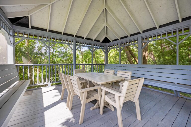 Wooden gazebo with a table, chairs, benches, and views of green trees through screened walls.