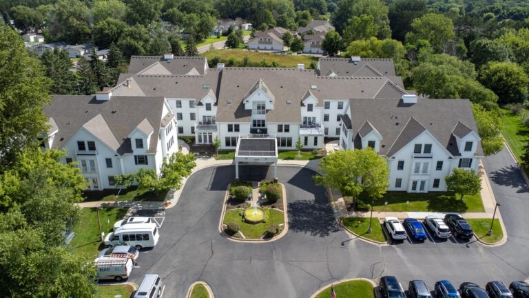 Aerial view of a large white residential building with a parking lot and trees surrounding it.