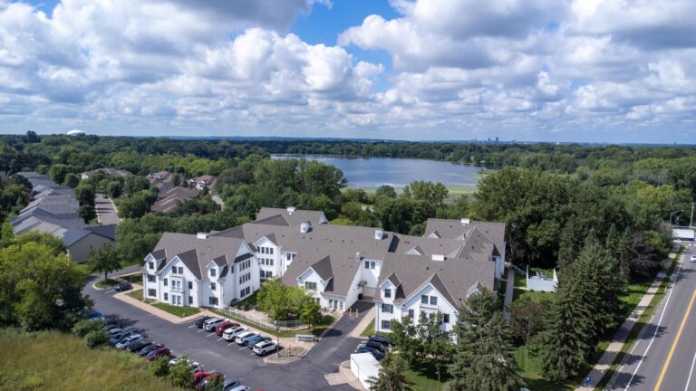 Aerial view of white apartment buildings near a lake, surrounded by trees and parked cars.