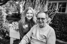 Smiling young woman stands behind an older man in a sweater, outdoors with flowers in the background. Black and white.