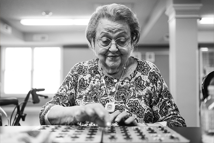 An older woman wearing glasses and a floral print blouse and emergency pendant sits at a table playing bingo.