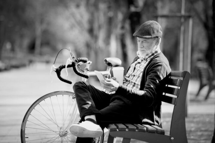 A senior man with bicycle in town park sitting on bench, holding coffee and smart phone, texting. Sunny spring day.