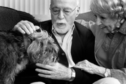 An older couple pets a dog that's standing on a sofa next to them. Black and white photo.