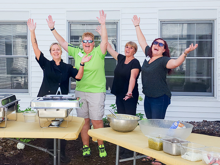 For female team members stand outside behind tables of food and raise their hands in celebration.